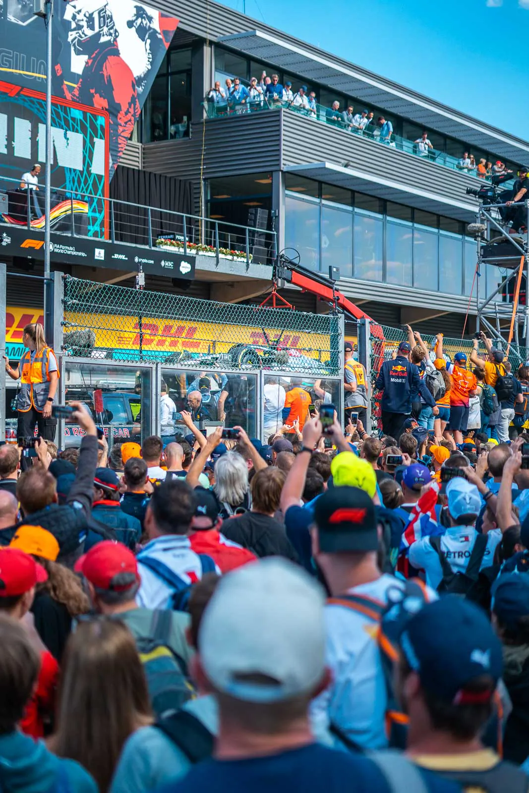 Fans mit erhobenen Handys vor einem Formel-1-Teamgebäude mit Zuschauern auf den Balkonen bei Sonnenschein.