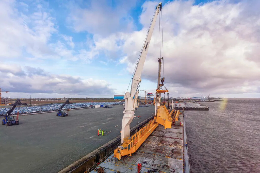Hafenkran hebt gelbes Industriebauteil über einem Frachtschiff in Cuxhaven, Arbeiter am Kai unter bewölktem Himmel.