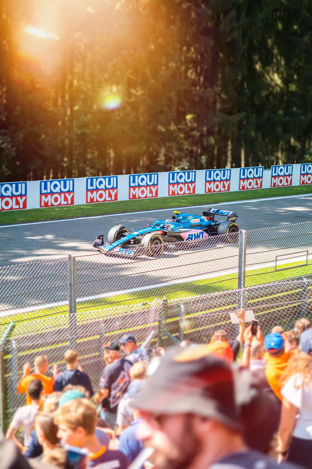 Blau-rosa Formel-1-Rennwagen auf der Strecke, Zuschauer hinter einem Zaun und warmes Sonnenlicht vor Waldkulisse.