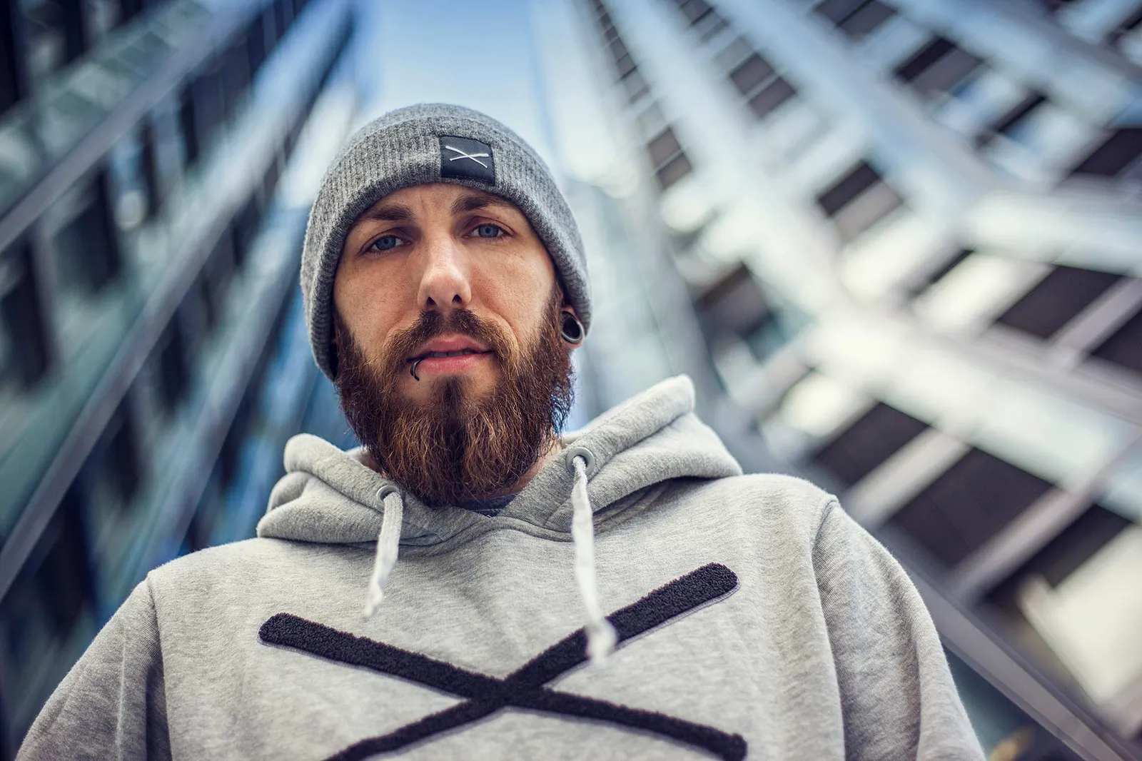 Bärtiger Mann mit grauer Mütze und Kapuzenpullover vor modernen Hochhausfassaden, aus der Untersicht bei Tageslicht.
