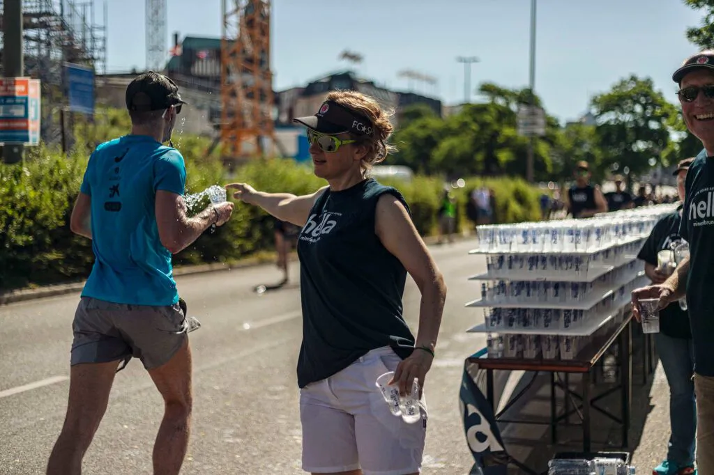 Helferin reicht einem Läufer bei Sonnenschein Wasser; daneben stehen Tische mit Trinkbechern am Straßenrand.
