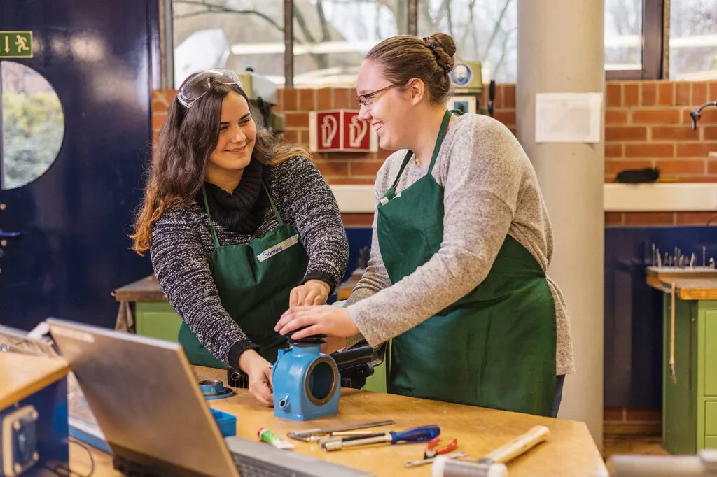 Zwei Frauen in grünen Schürzen arbeiten lächelnd an einem blauen Bauteil auf einer Werkbank in einer hellen Werkstatt.