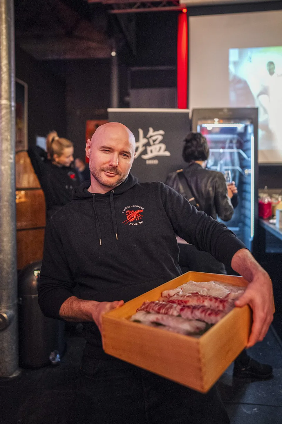 Mann im schwarzen Hoodie hält eine Holzkiste mit Fisch auf Eis; im Hintergrund Gäste und ein Kühlgerät beim Event.