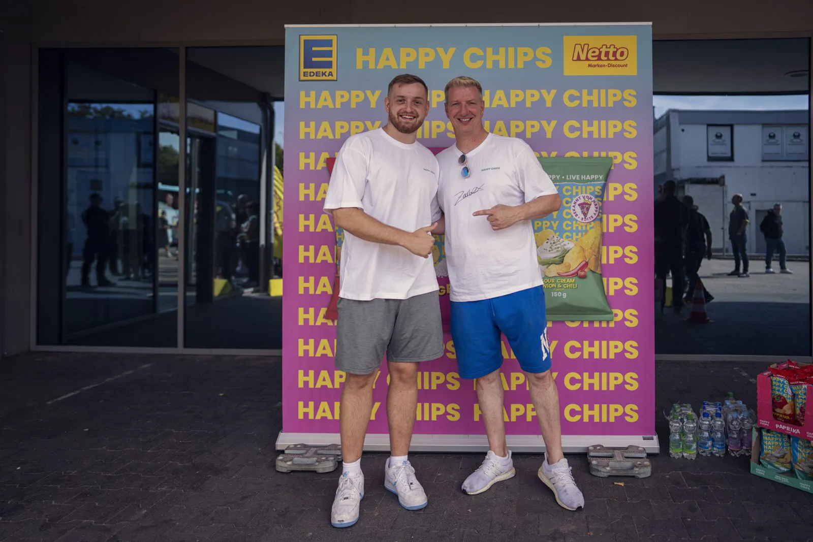 Zwei Männer in weißen Shirts posieren vor einer Netto-Happy-Chips-Wand am Eingang mit Glastüren.