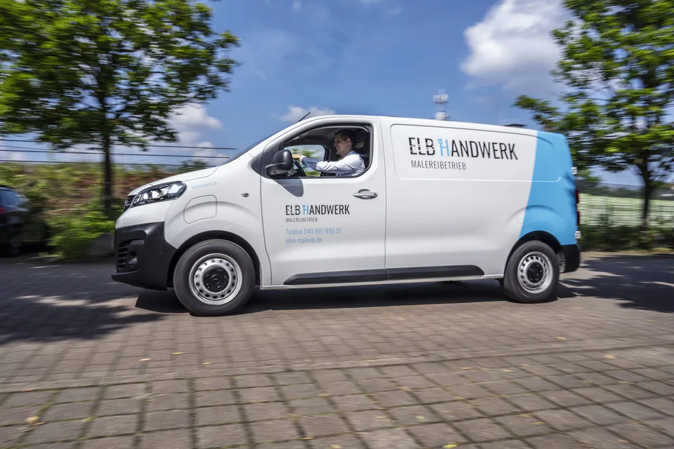 Fahrer im weiß-blauen Transporter von Elb Handwerk auf einer sonnigen, baumgesäumten Straße.