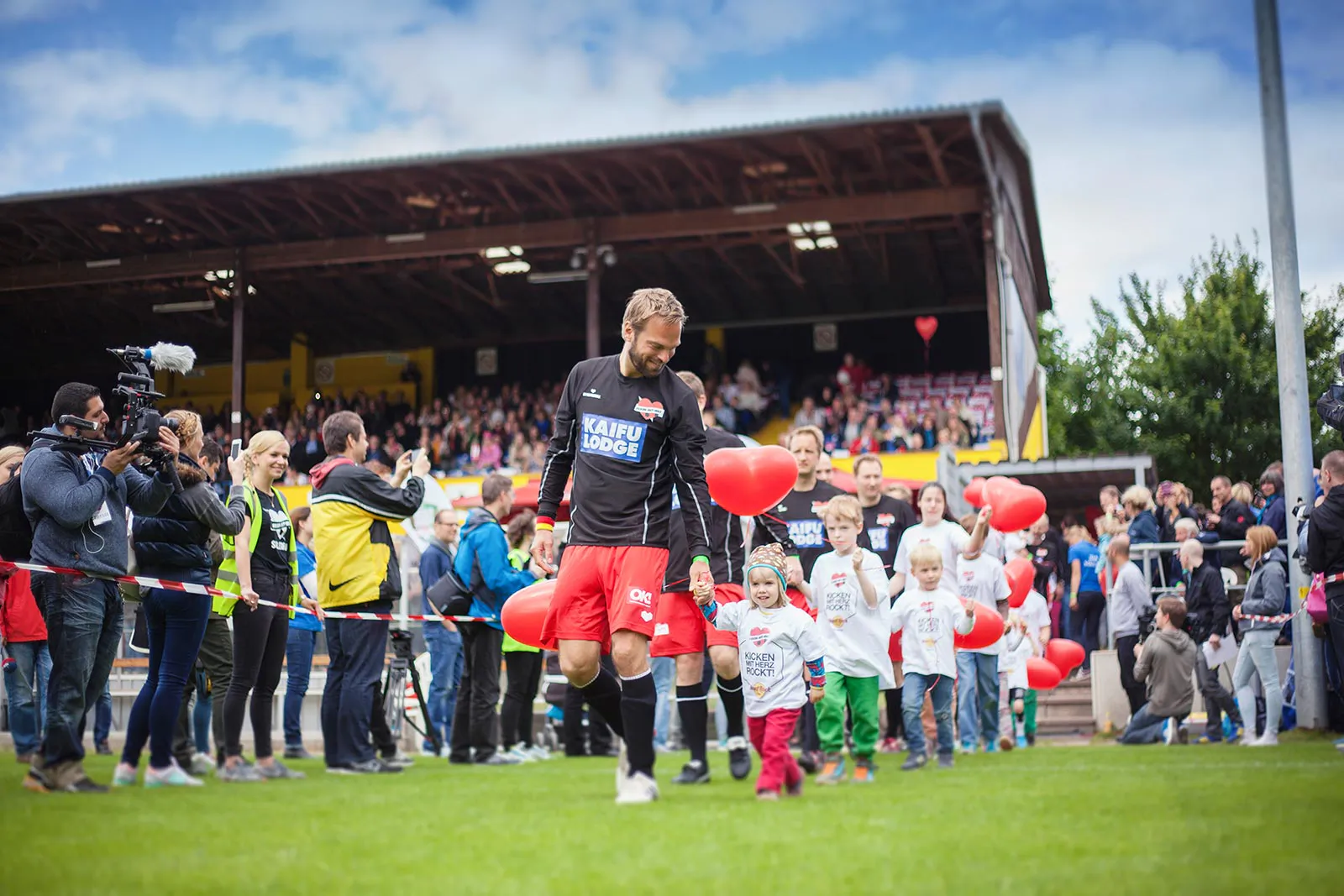 Fußballspieler und Kinder mit roten Herzballons laufen bei Tageslicht über den Rasen vor einer besetzten Tribüne.