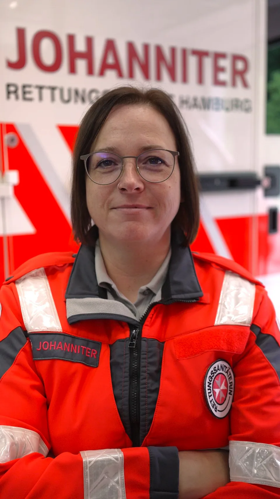 Frau mit Brille in roter Johanniter-Rettungsdienstjacke steht mit verschränkten Armen vor einem Rettungswagen.