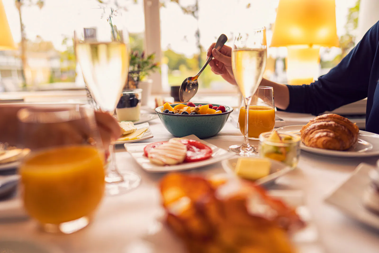 Person mit Löffel über einer Obstschale am sonnigen Frühstückstisch mit Croissants, Orangensaft und Sektgläsern.