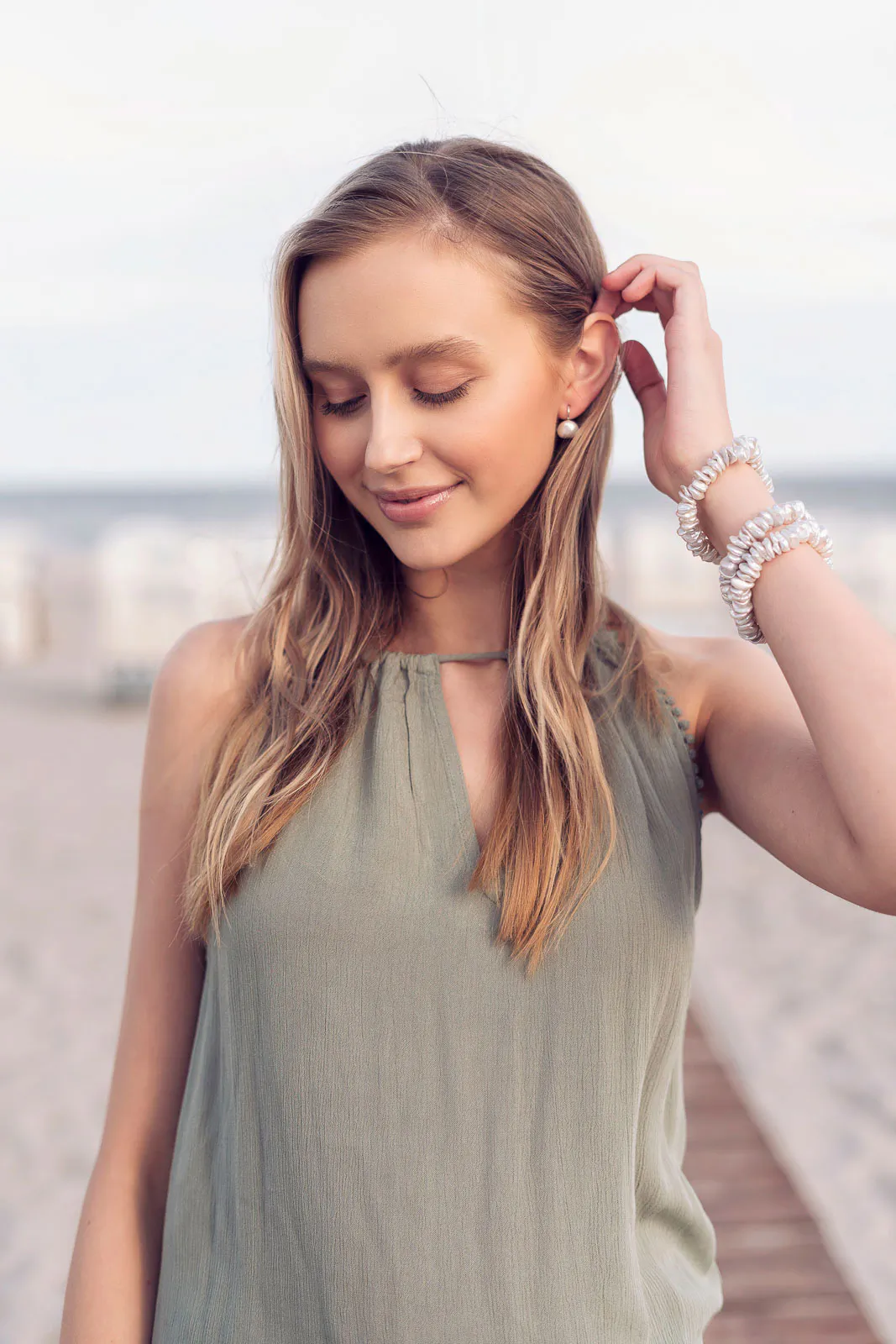 Frau im olivgrünen Kleid mit Perlenarmbändern und -ohrringen auf einem Strandsteg bei weichem Tageslicht.