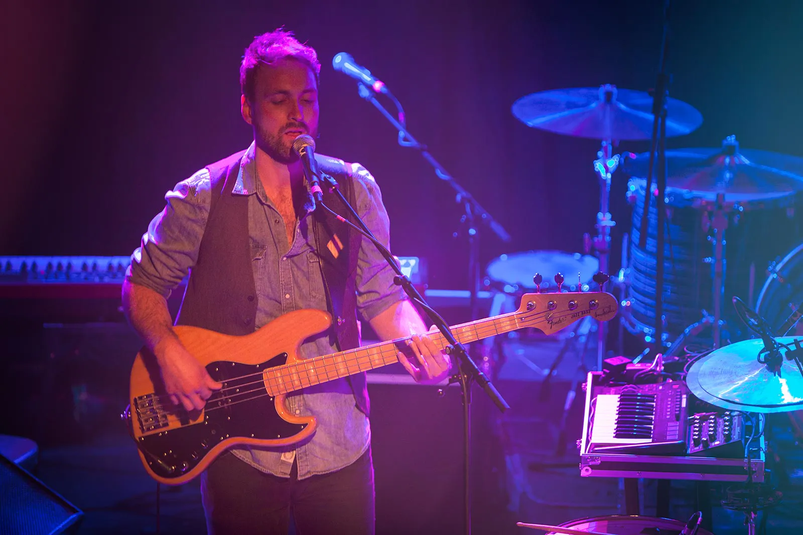 Bassist singt am Mikrofon auf einer violett-blau beleuchteten Konzertbühne, dahinter Schlagzeug und Keyboard.