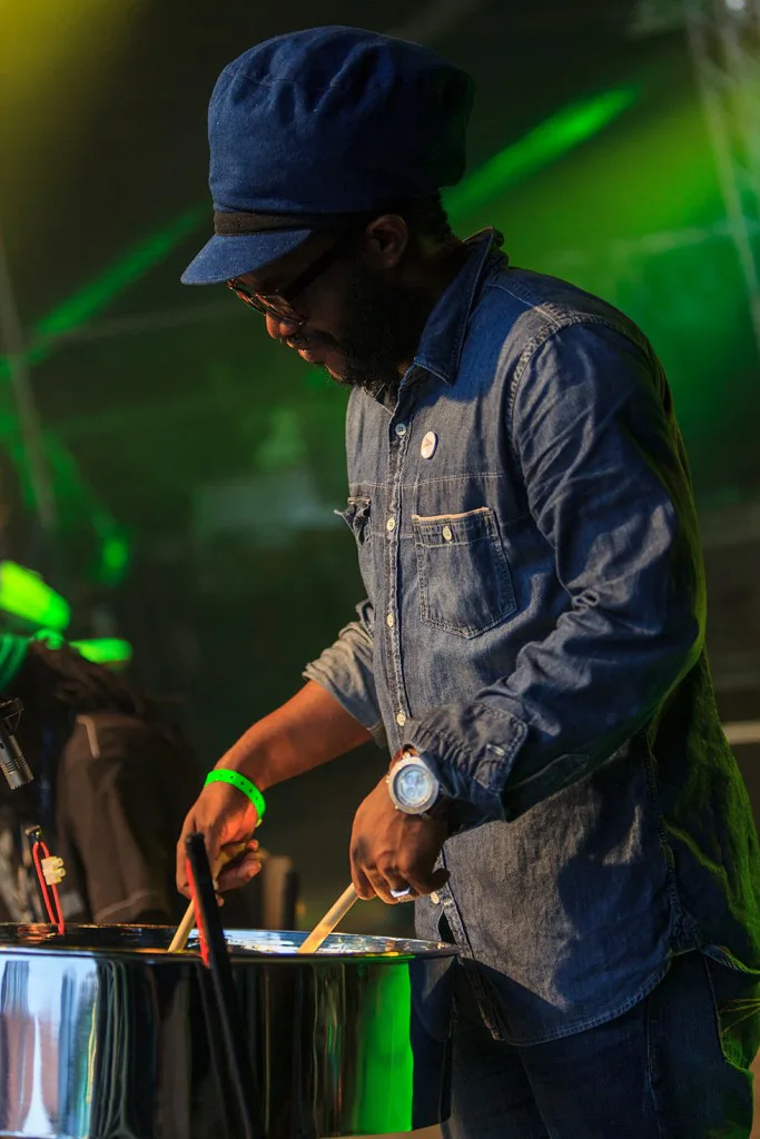 Musiker mit blauer Mütze und Jeanshemd spielt eine Steelpan auf einer grün beleuchteten Bühne.