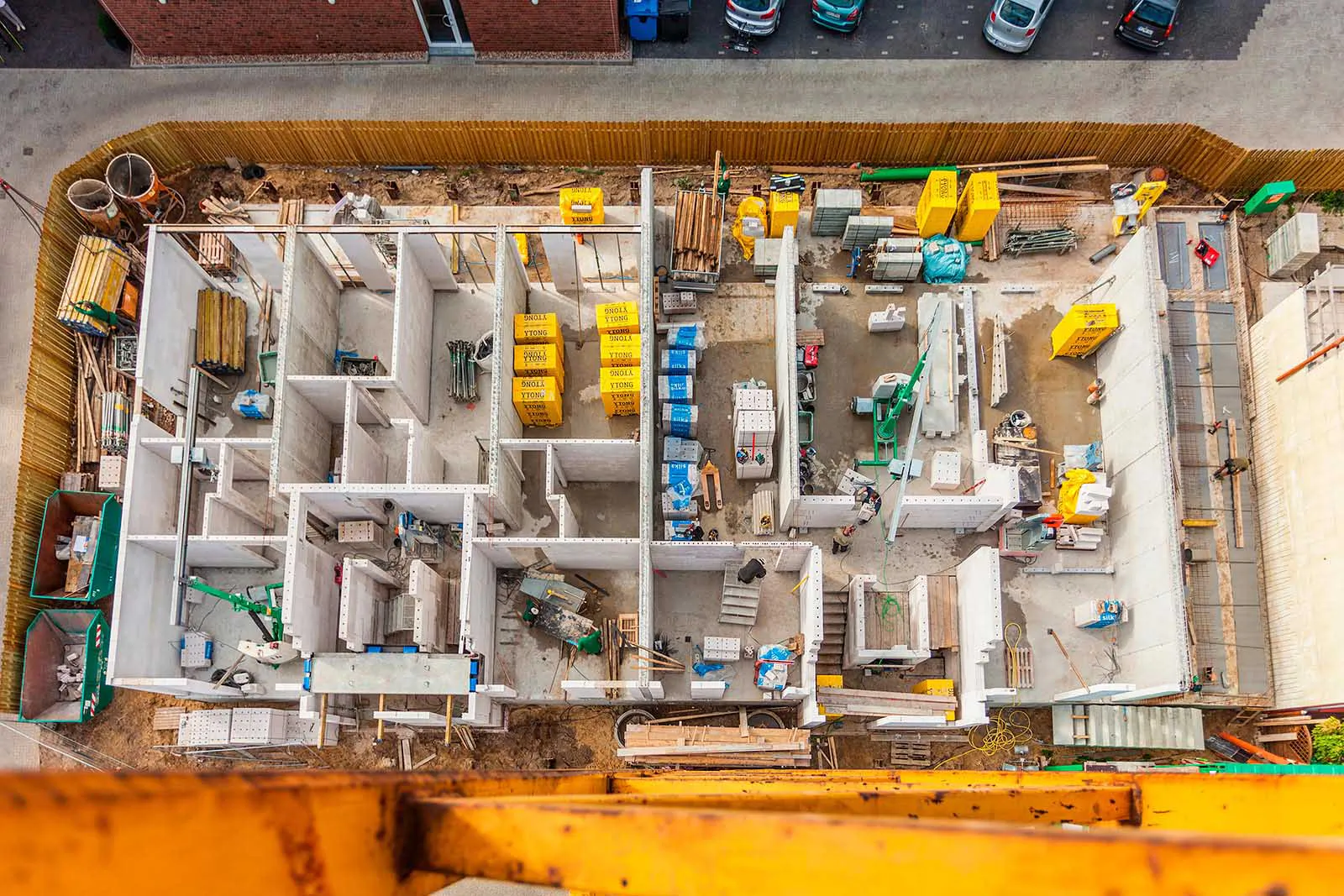 Blick von oben auf eine Baustelle mit offenen Rohbauräumen, Baumaterial und gelbem Kranträger im Vordergrund.