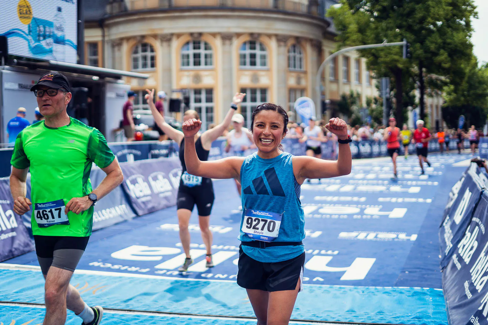 Lächelnde Läuferin im blauen Trikot jubelt mit erhobenen Armen im Zielbereich des Hella Halbmarathons.