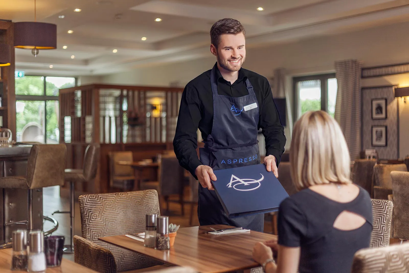 Lächelnder Kellner mit Schürze reicht einer Frau am Holztisch eine Speisekarte im warm beleuchteten Restaurant.