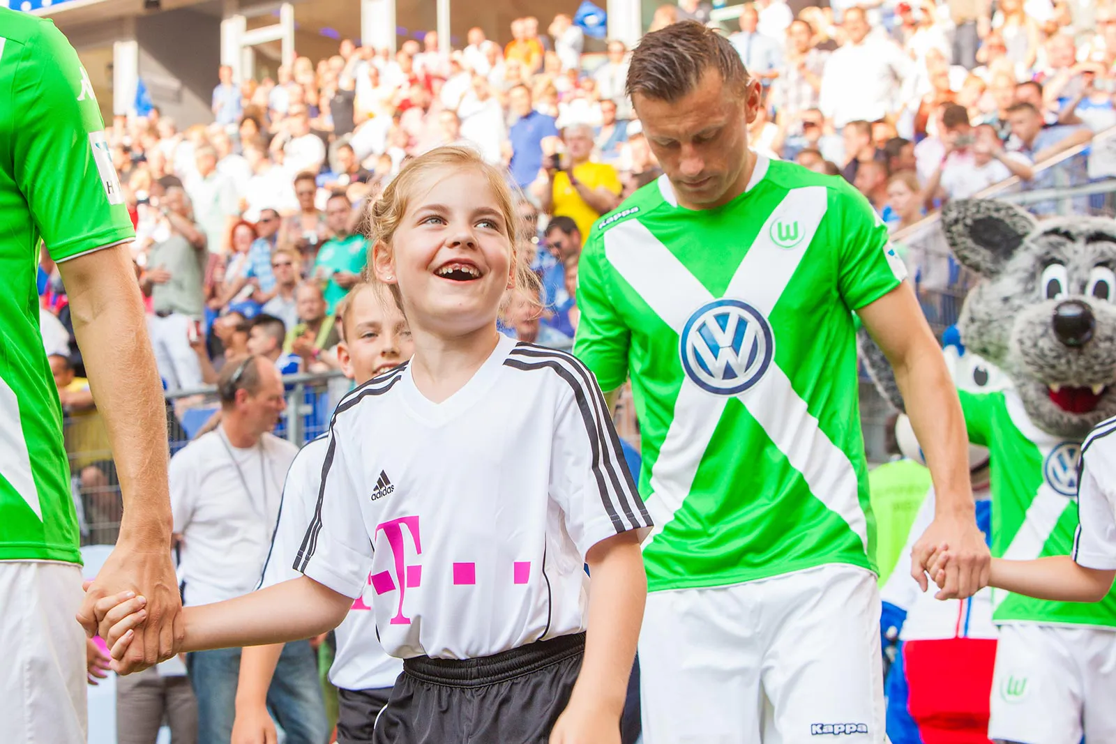 Lachendes Kind im weißen Trikot hält die Hand eines Fußballspielers; volle Tribüne und Maskottchen im Sonnenlicht.