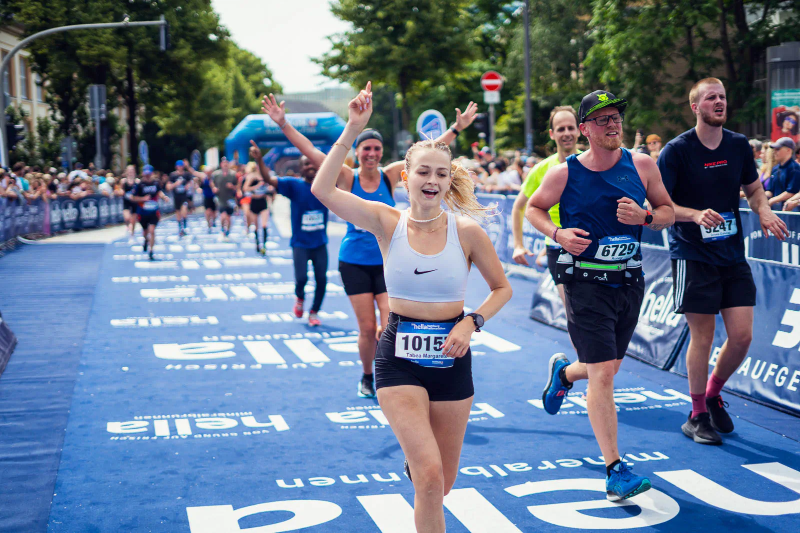 Jubelnde Läuferin mit erhobenem Arm auf blauem Teppich beim Hella Halbmarathon, umgeben von weiteren Läufern.