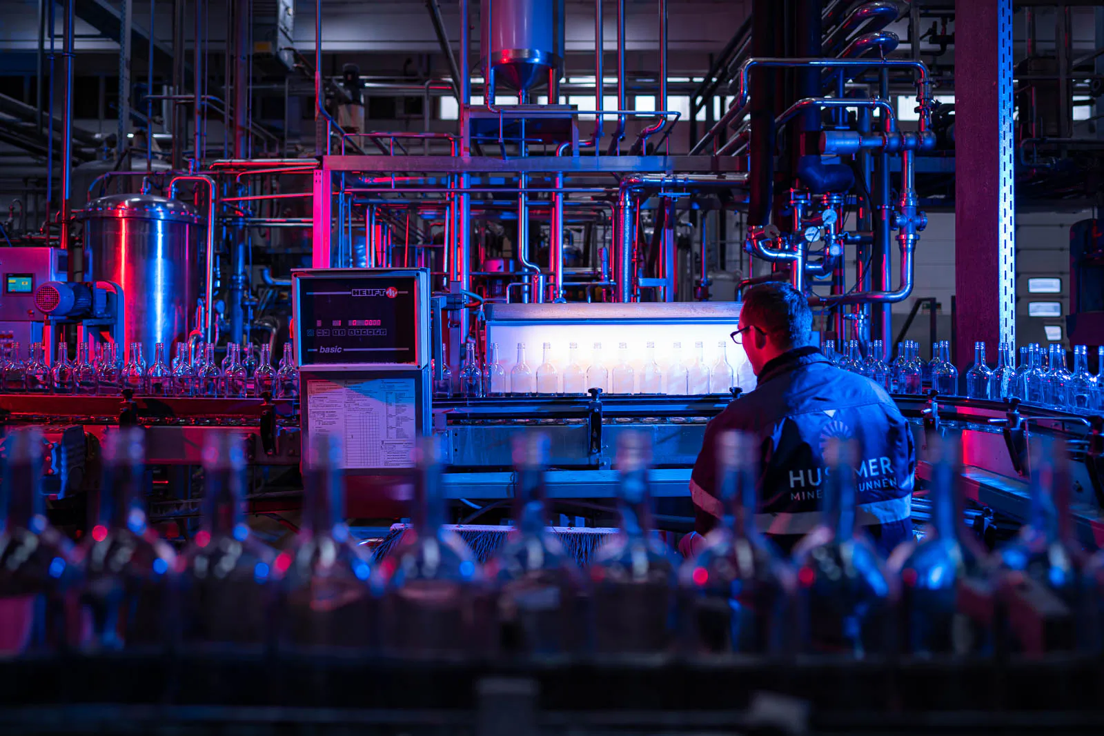 Mitarbeiter an der Abfüllanlage von Husumer Mineralbrunnen, umgeben von Glasflaschen in blau-rotem Licht.
