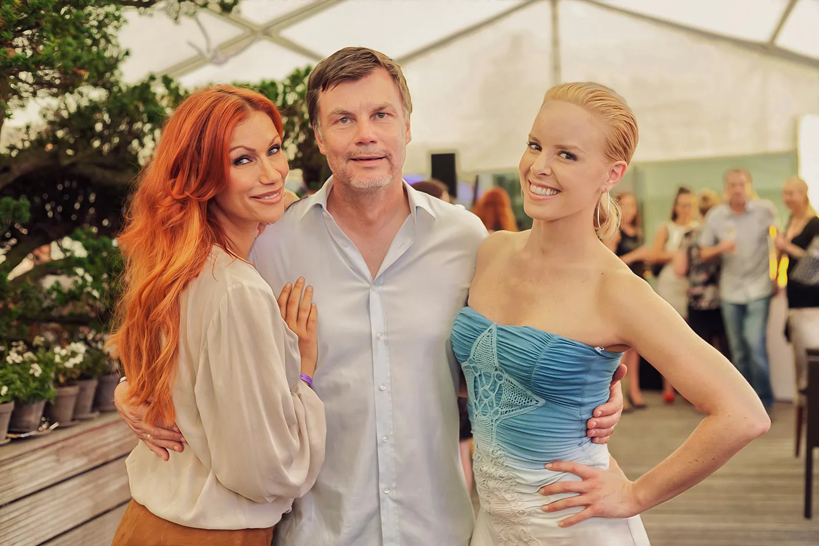 Zwei Frauen und ein Mann posieren eng beieinander im hellen Festzelt, dahinter Partygäste und Pflanzen.