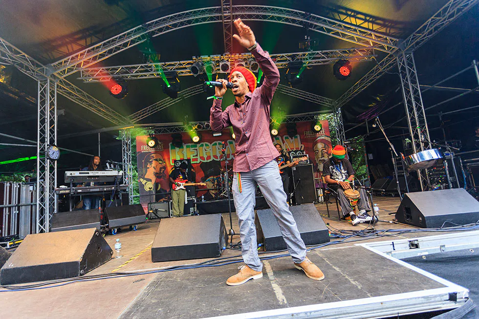 Sänger mit bunter Mütze und Mikrofon hebt den Arm auf einer überdachten Bühne, dahinter Musiker und farbiges Licht.