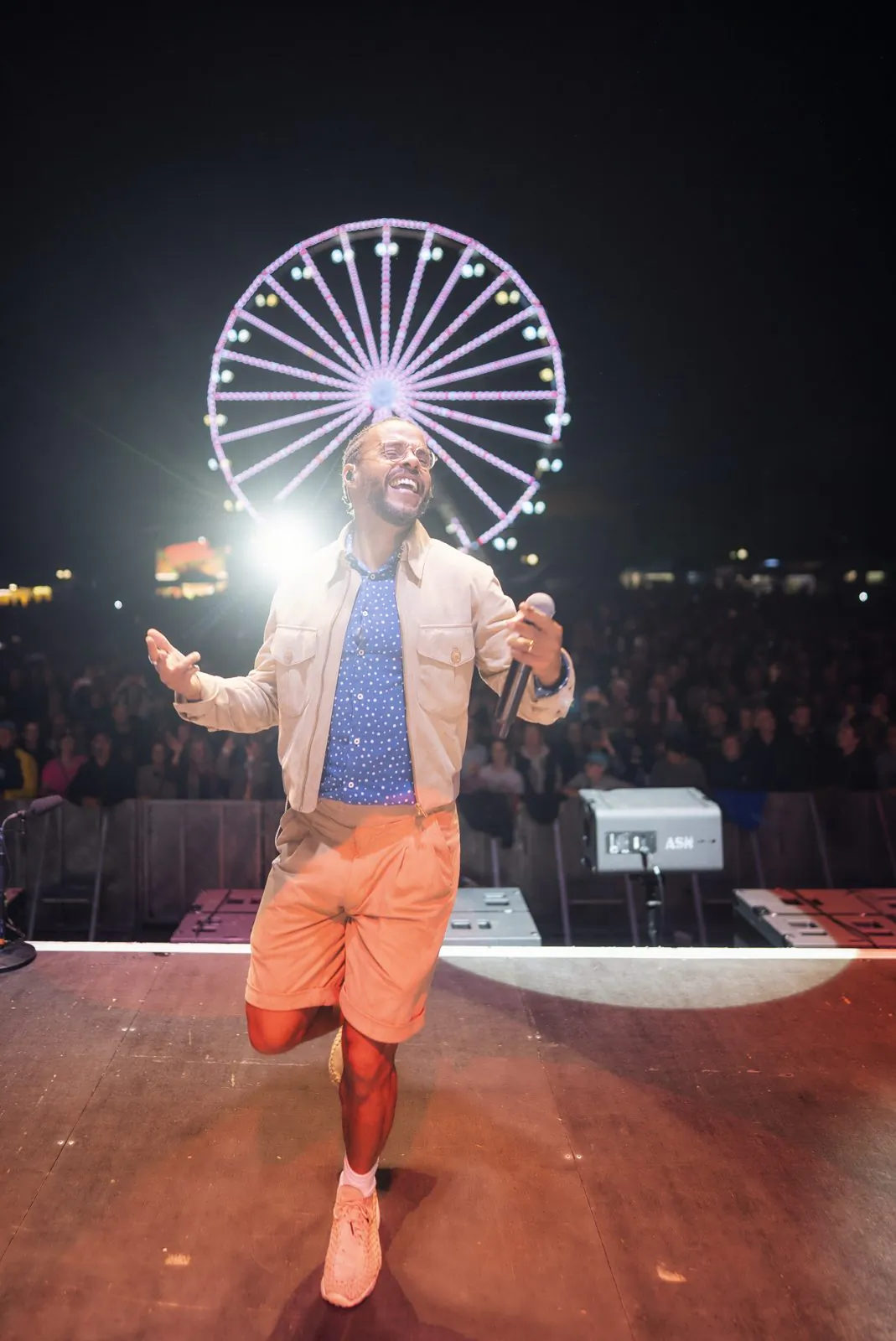 Sänger mit Mikrofon und orangefarbenen Shorts auf der Bühne, dahinter Publikum und ein leuchtendes Riesenrad bei Nacht.