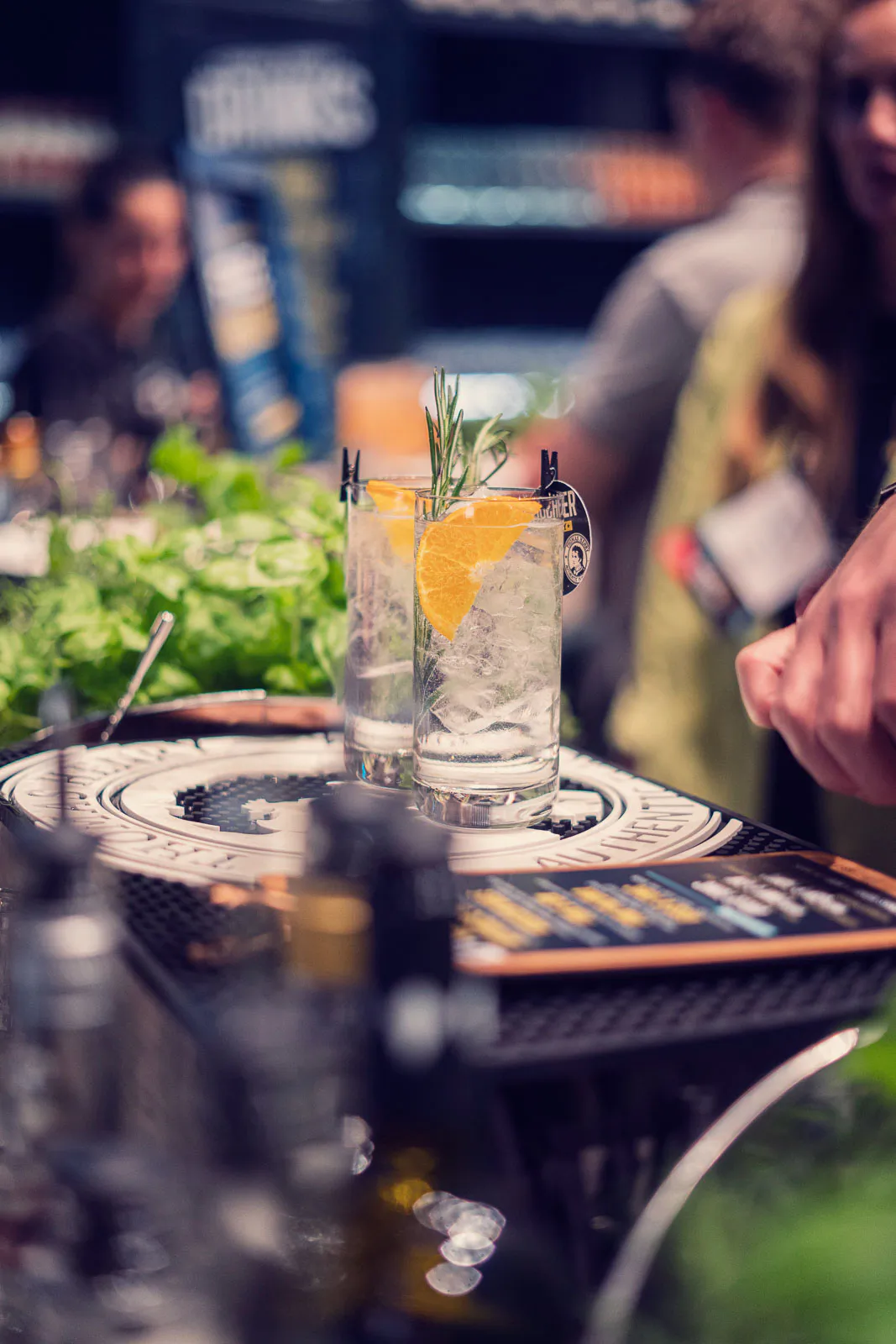 Zwei Longdrinks mit Eis, Orangenschale und Rosmarin auf einer Thomas-Henry-Bartheke, dahinter unscharfe Gäste.