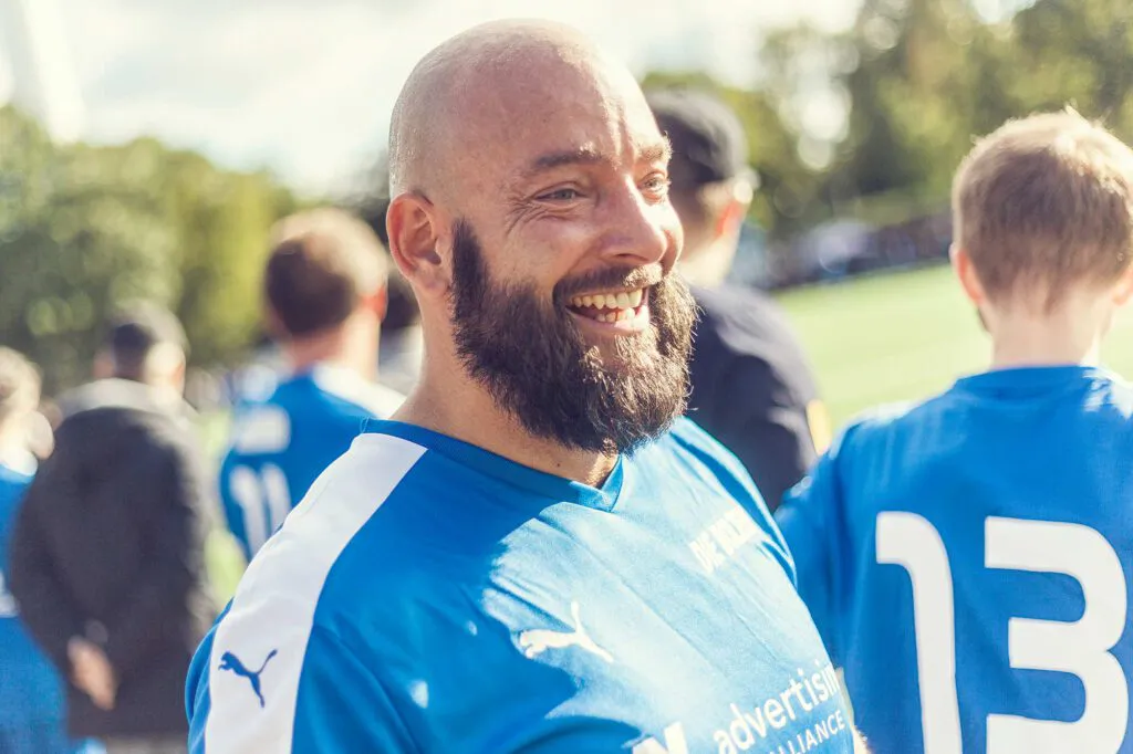 Lachender Mann mit Glatze und Bart im blauen Fußballtrikot, umgeben von Spielern auf einem sonnigen Fußballplatz.