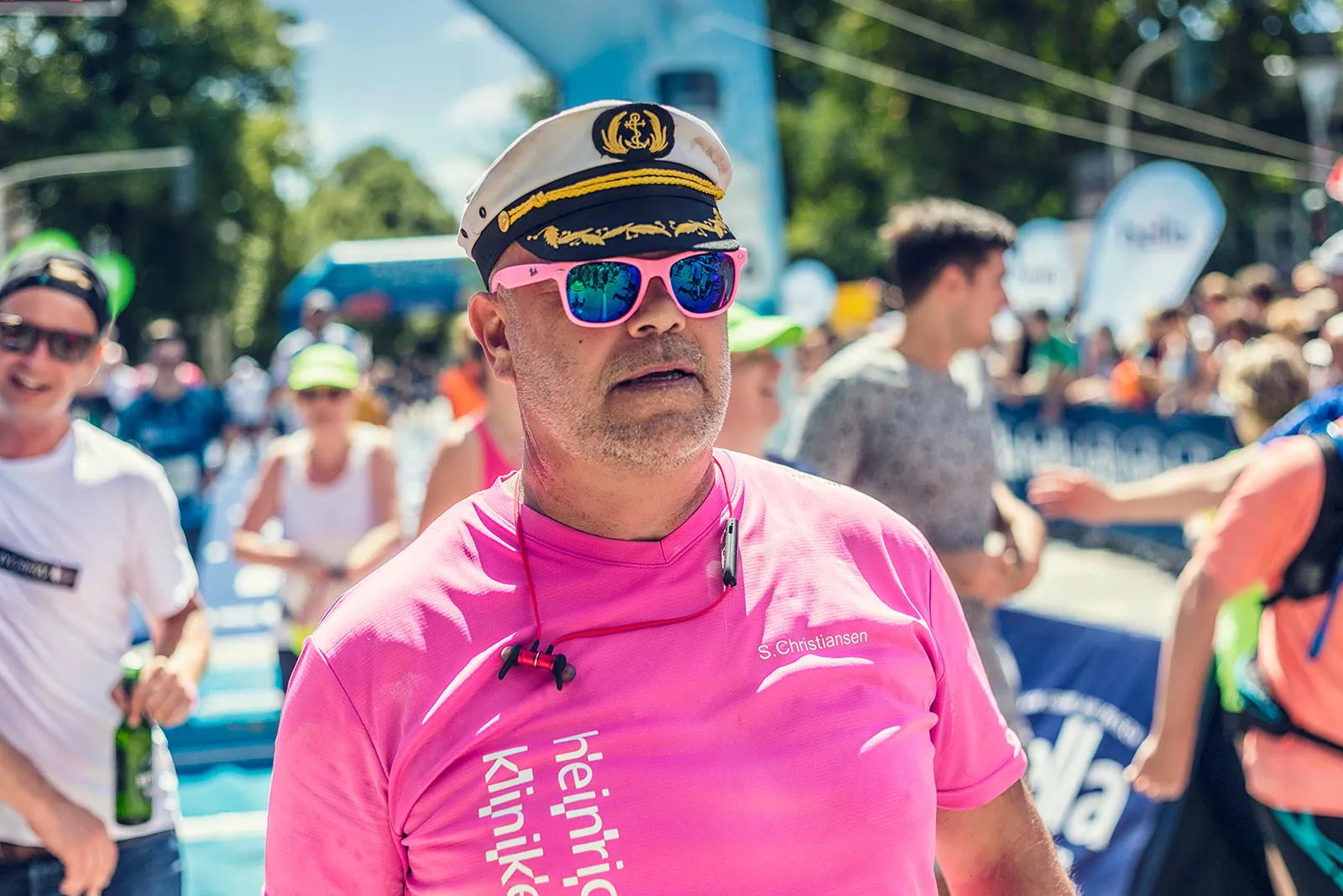 Mann mit Kapitänsmütze, pinker Sonnenbrille und pinkem Laufshirt zwischen Teilnehmenden auf sonniger Straße.