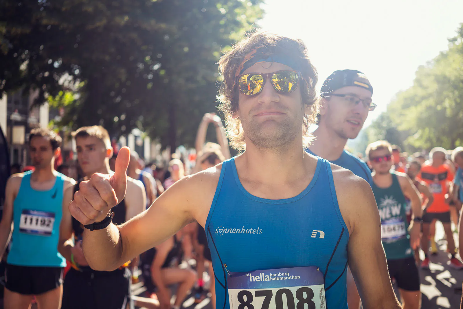 Läufer mit Sonnenbrille und blauem Trikot zeigt den Daumen nach oben, umgeben von Läufern auf sonniger Straße.