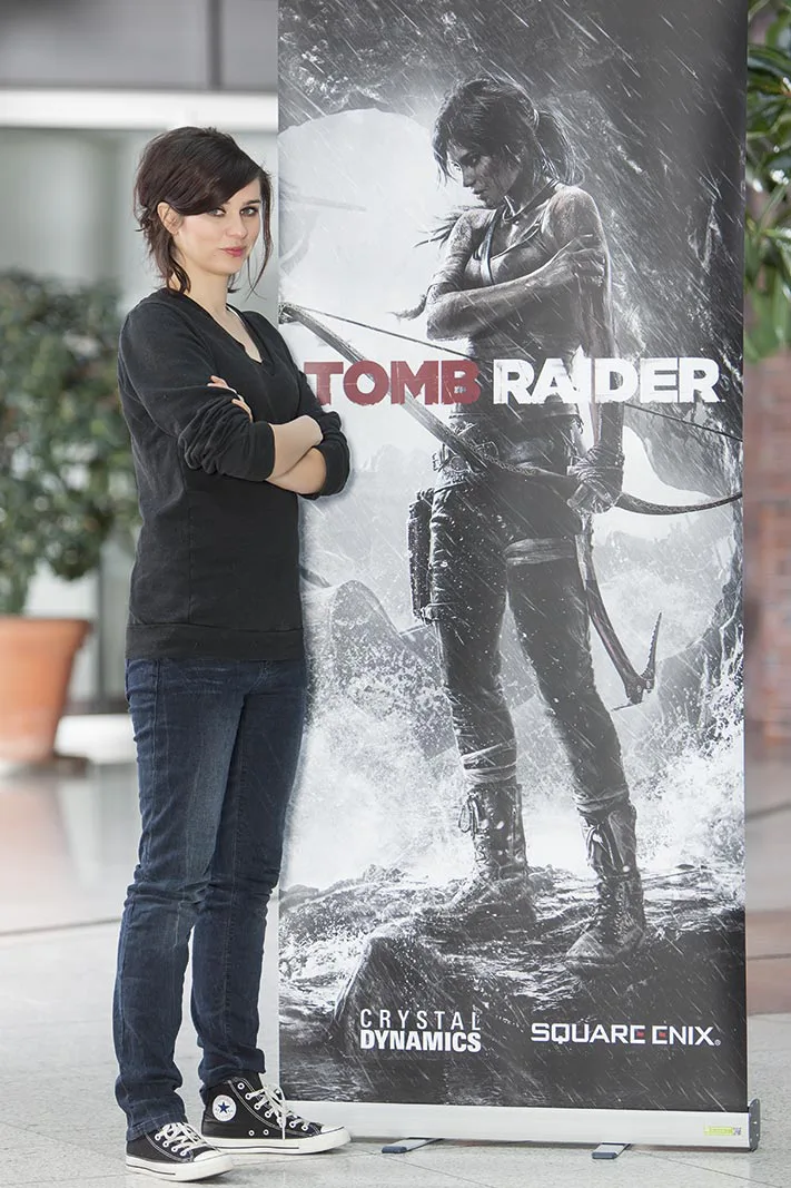 Nora Tschirner steht mit verschränkten Armen neben einem Tomb-Raider-Aufsteller in einem hellen Innenraum.