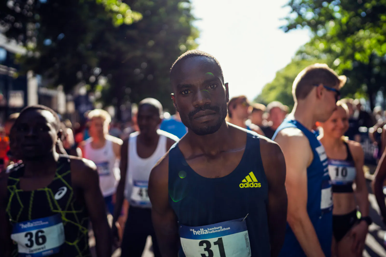 Läufer mit Startnummer 31 im dunklen Adidas-Trikot steht zwischen weiteren Teilnehmern auf sonniger, baumgesäumter Straße.