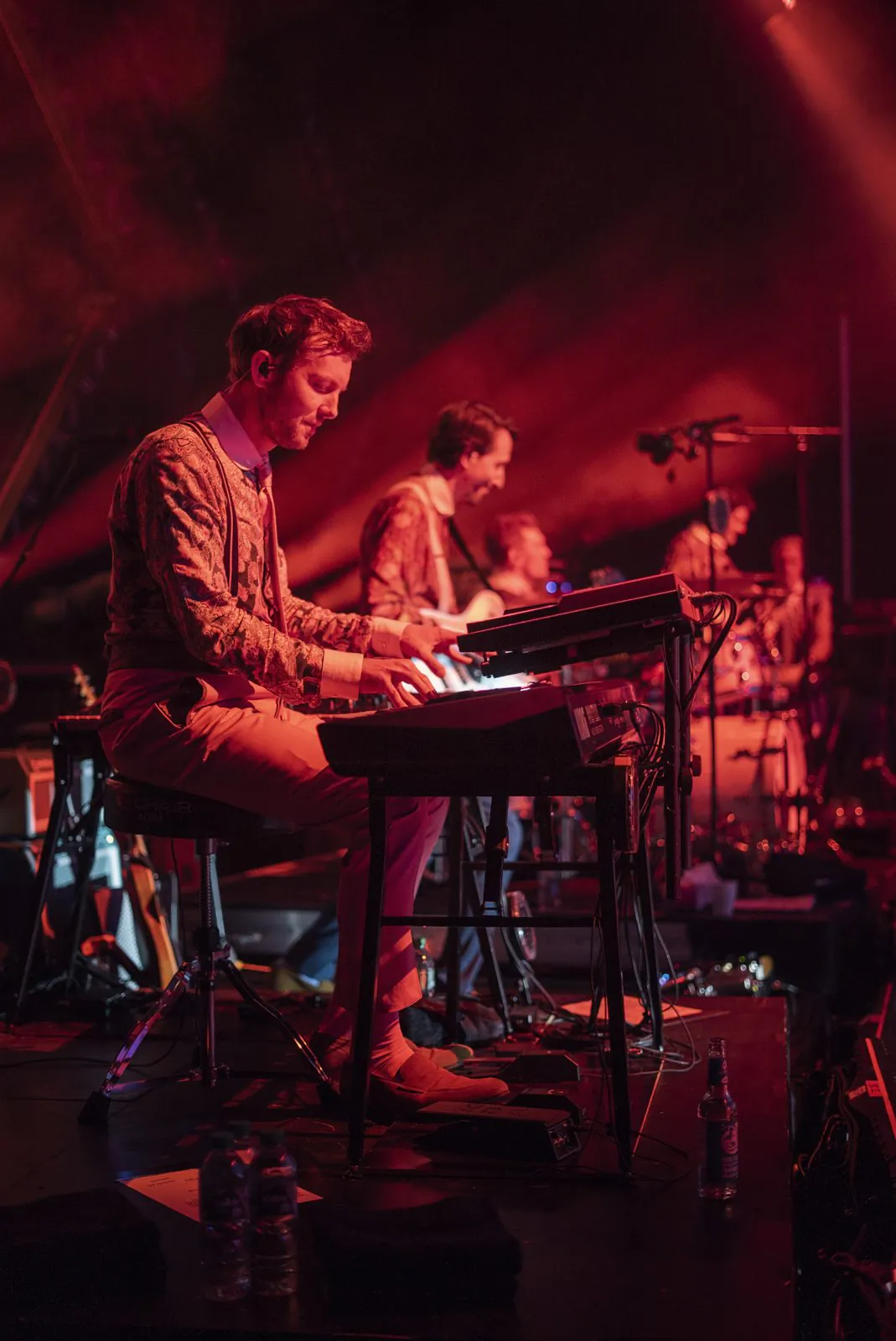 Keyboarder spielt auf einer rot beleuchteten Konzertbühne, weitere Musiker stehen im Hintergrund.