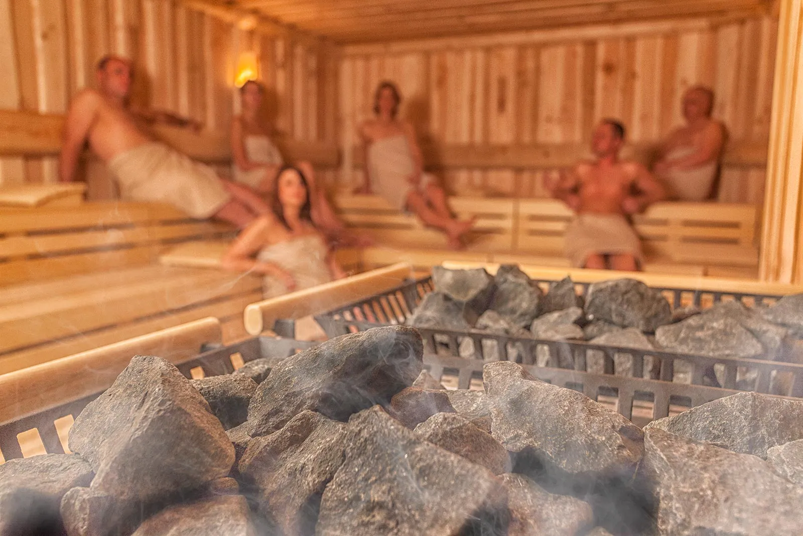 Saunasteine im Vordergrund, sechs Personen mit Handtüchern auf Holzbänken einer warm beleuchteten Sauna.