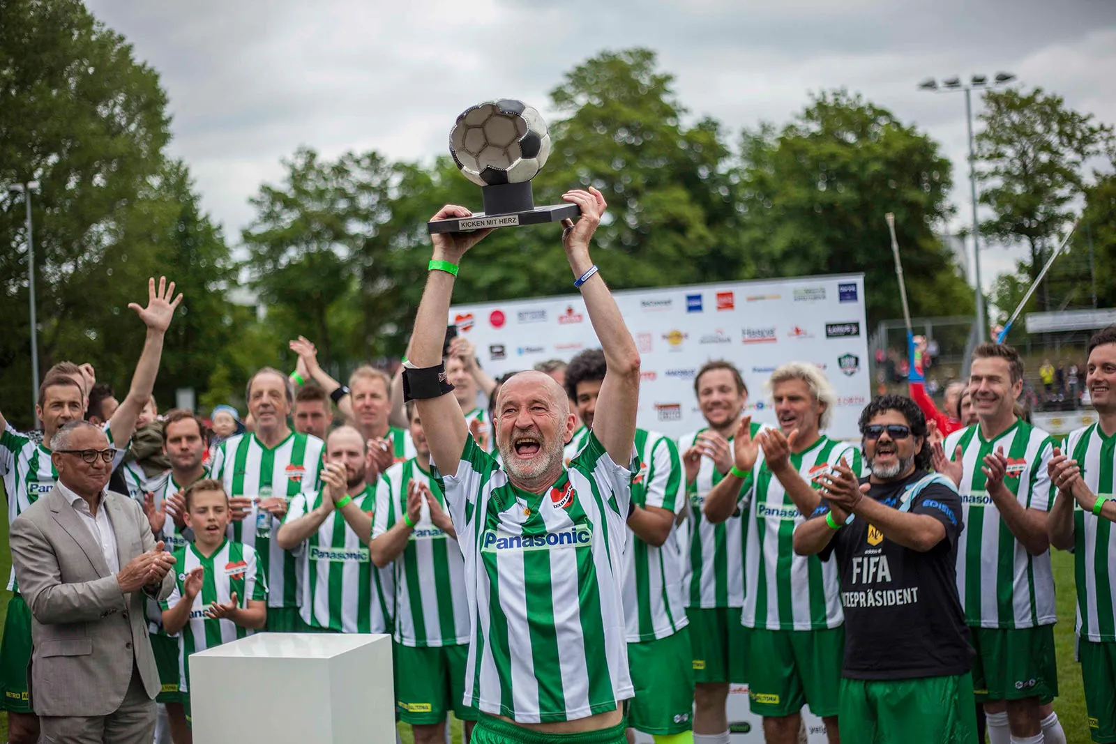 Jubelnder Spieler hebt einen Fußballpokal, umringt von klatschenden Männern in grün-weißen Trikots auf dem Sportplatz.