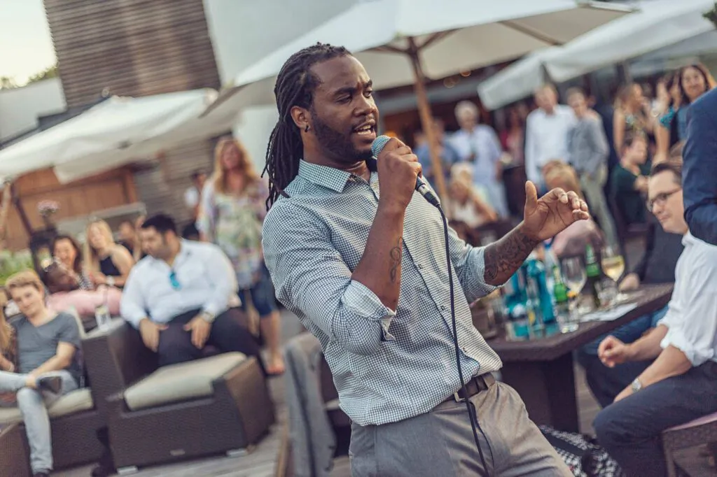 Sänger mit Dreadlocks singt ins Mikrofon zwischen Gästen auf einer sonnigen Terrasse mit Lounge-Möbeln und Sonnenschirmen.
