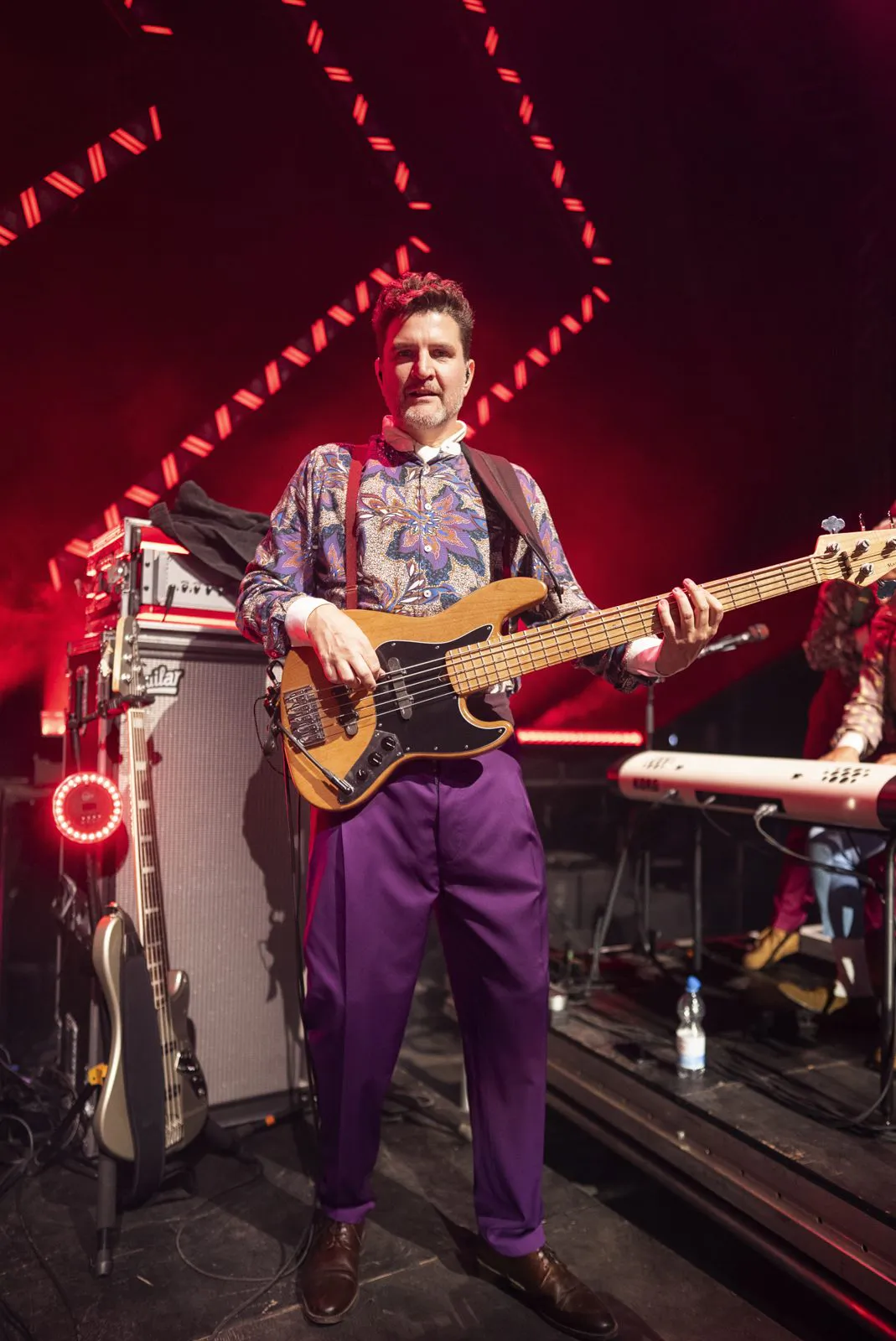 Bassist in gemustertem Hemd und lila Hose spielt E-Bass auf rot beleuchteter Bühne neben Verstärker und Keyboard.