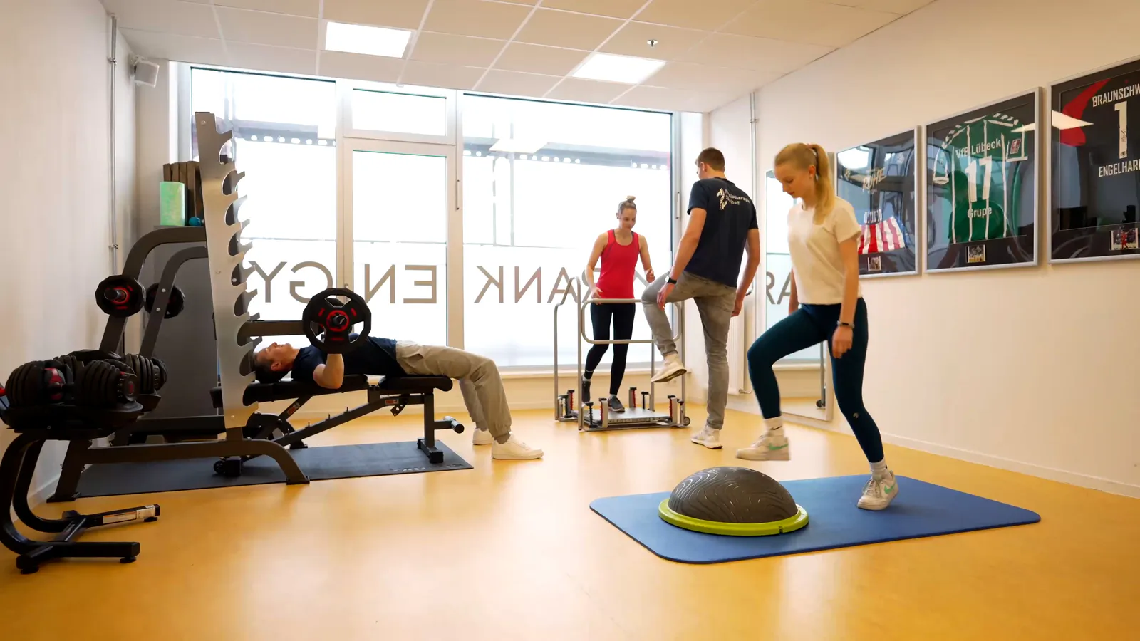 Vier Personen trainieren im hellen Physiotherapieraum mit Hantelbank, Stepper und Balancegerät.