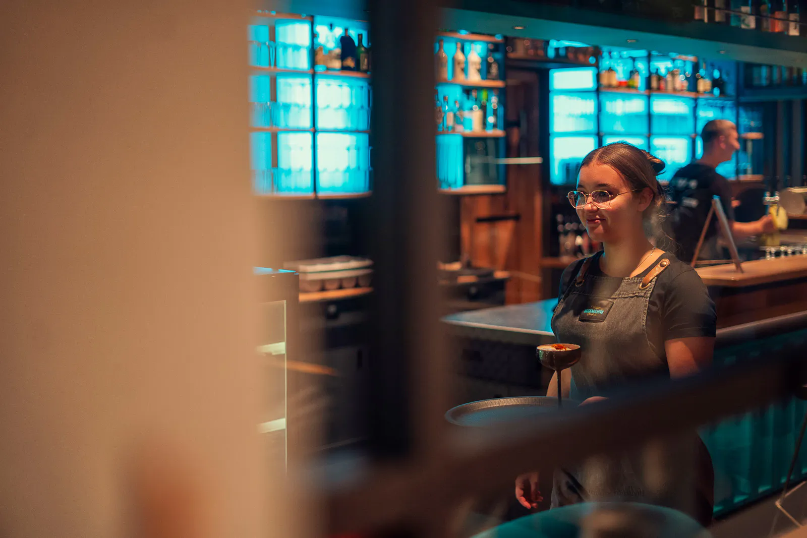 Lächelnde Servicekraft vor der türkis beleuchteten Bar in Beckmanns Strandhalle, im Vordergrund unscharfe Holzlamellen.