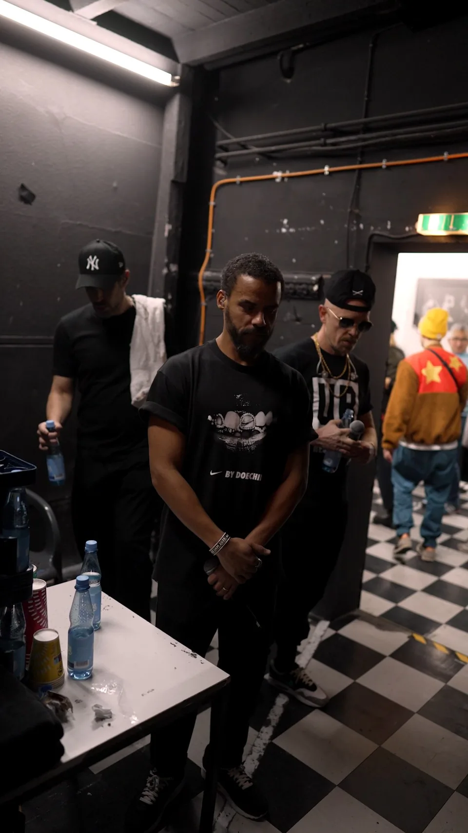 Mehrere Männer stehen in einem dunklen Backstage-Raum mit Neonlicht, Getränketisch und schwarz-weiß kariertem Boden.