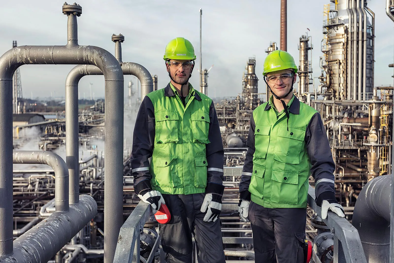 Zwei Personen mit grünen Helmen und Schutzkleidung auf einer Metalltreppe zwischen Rohrleitungen der Raffinerie Heide.