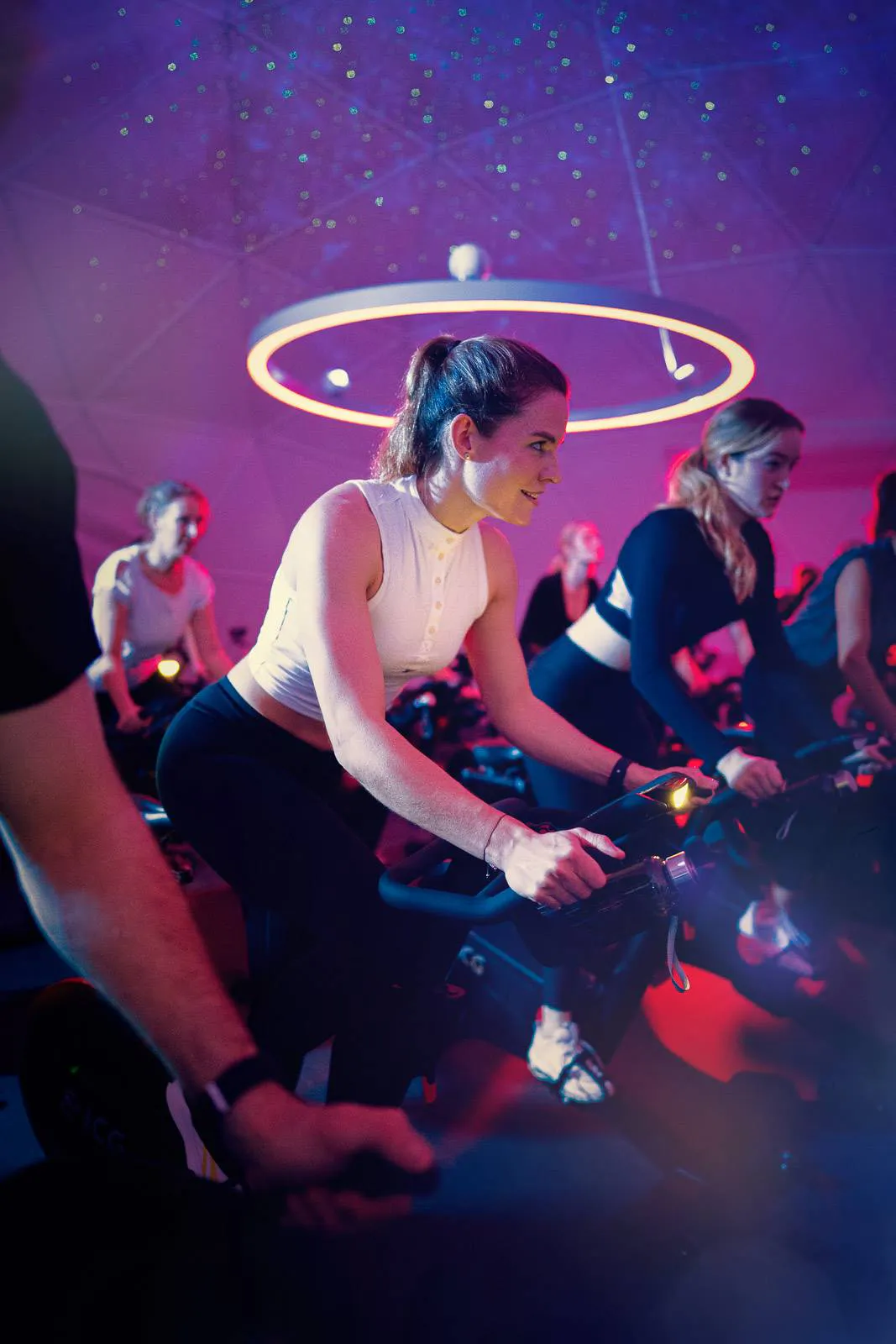 Frauen trainieren auf Spinningrädern im Fitnessstudio unter violett-rotem Licht und einer ringförmigen Deckenleuchte.