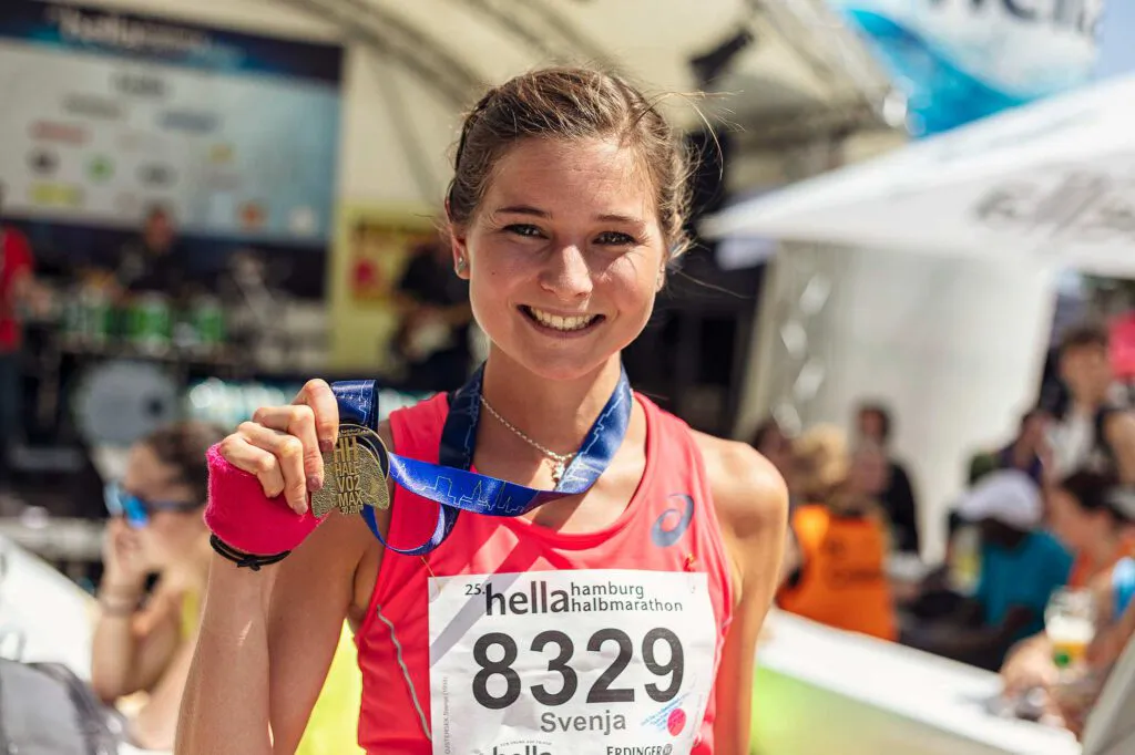 Lächelnde Läuferin im roten Top mit Startnummer 8329 hält ihre Medaille im Sonnenlicht vor Veranstaltungszelten.
