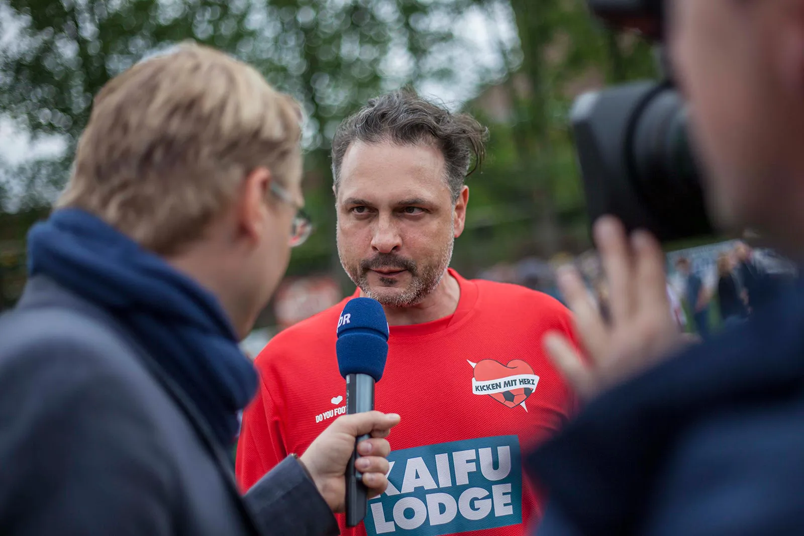 Mann im roten Kicken-mit-Herz-Shirt gibt draußen ein Interview mit blauem Mikrofon, rechts eine Kamera.