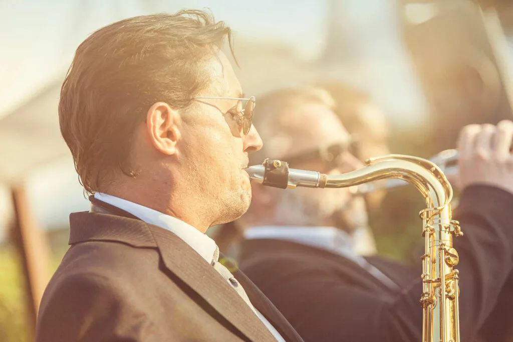 Saxofonist im Anzug mit Sonnenbrille spielt im warmen Sonnenlicht, dahinter ein weiterer Bläser.