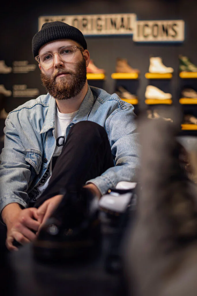 Bärtiger Mitarbeiter mit Brille, Mütze und Jeansjacke sitzt im Dr. Martens-Laden vor beleuchteten Schuhregalen.