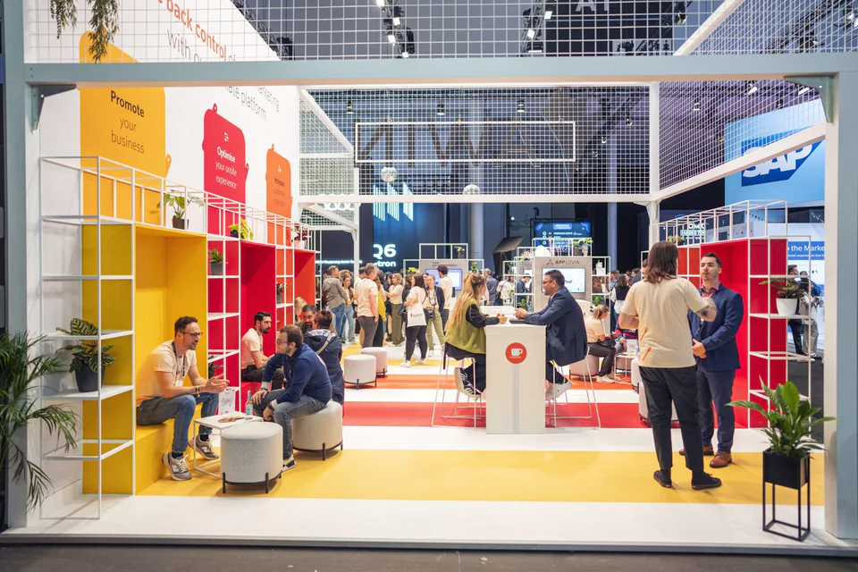 Besucher sitzen und unterhalten sich am hell beleuchteten Awin-Messestand mit gelben und roten Regalen.