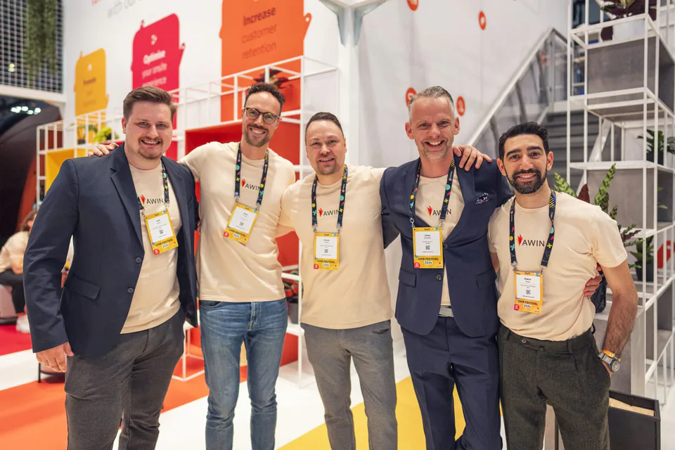 Fünf Männer in Awin-Shirts und mit Messeausweisen posieren lächelnd Arm in Arm vor einem hell beleuchteten Messestand.