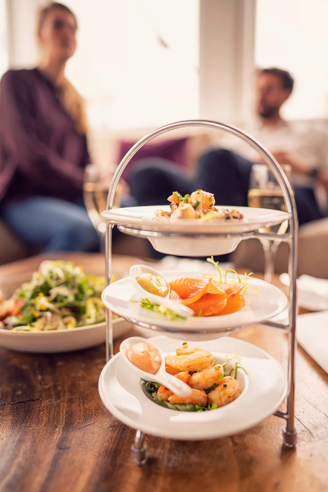 Dreistöckige Etagere mit Garnelen, Lachs und Häppchen auf einem Holztisch, zwei Gäste unscharf im hellen Hintergrund.