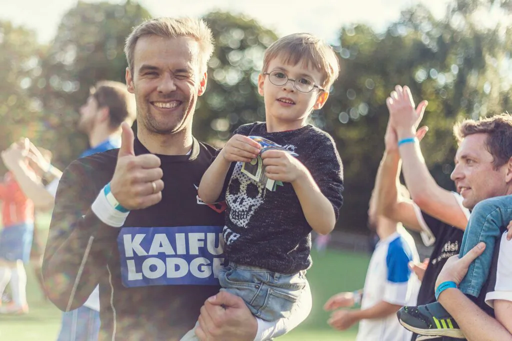 Lächelnder Mann hält einen Jungen auf dem Arm und zeigt den Daumen hoch, im Sonnenlicht auf einem Fußballplatz.