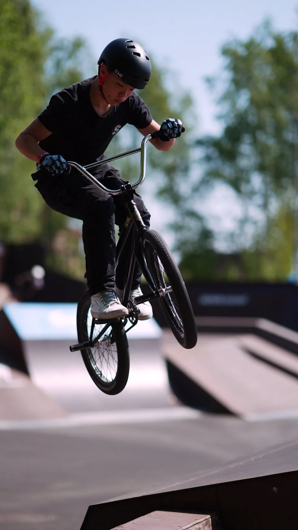 BMX-Fahrer mit schwarzem Helm springt bei Sonnenschein über eine Rampe im Skatepark, im Hintergrund Bäume.