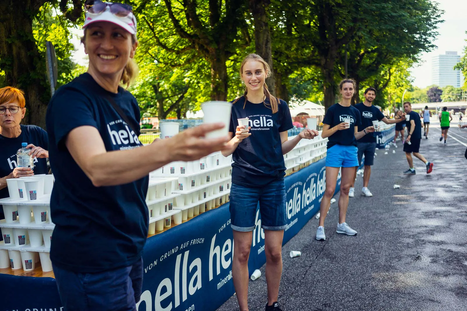 Helfer reichen Becher an einer Verpflegungsstation des Hella Halbmarathons auf einer sonnigen, baumgesäumten Straße.